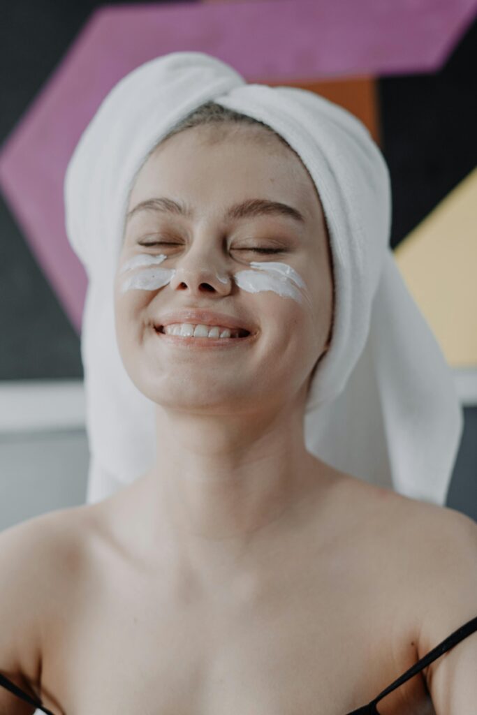 Smiling young woman with towel and face cream enjoying skincare routine.